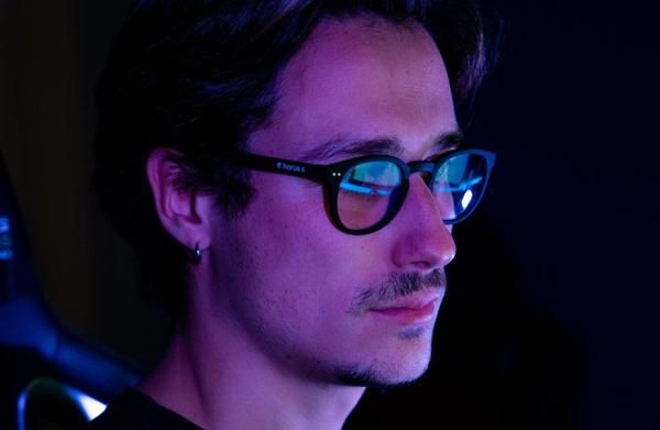Homme portant des lunettes rondes noires dans la pénombre, reflets d'écran bleus sur les verres